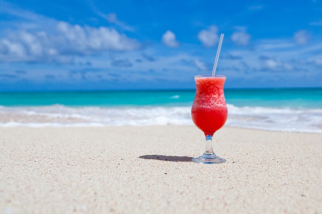 Red icy cocktail on a yellow sand beach with a green water and blue sky in the background